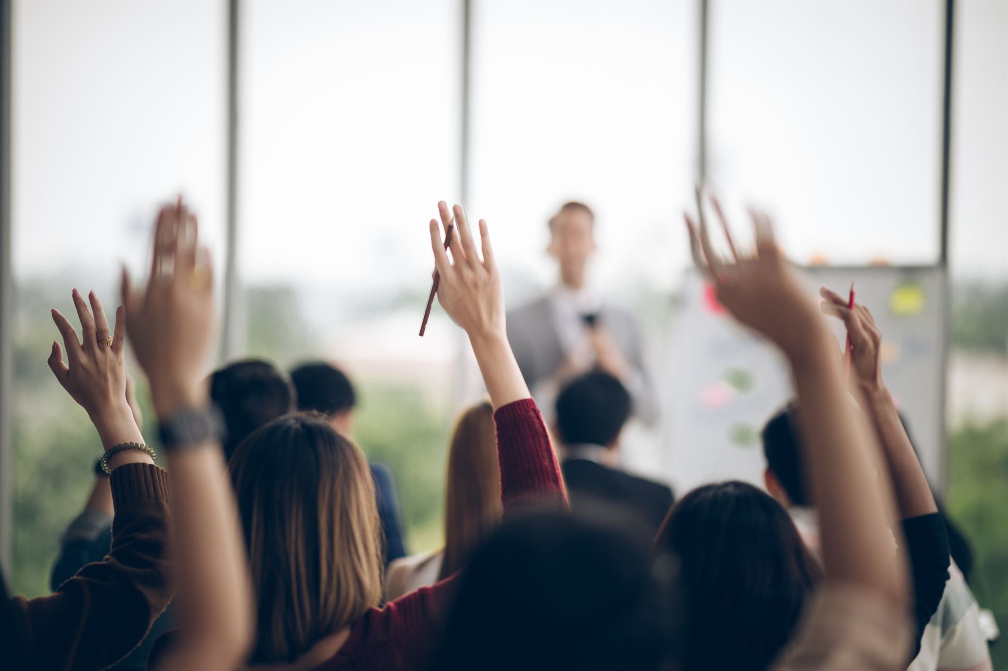 Back-of-peoples-heads-and-raised-hands-at-corporate-presentation-with-speaker-and-whiteboard-out-of-focus-in-background